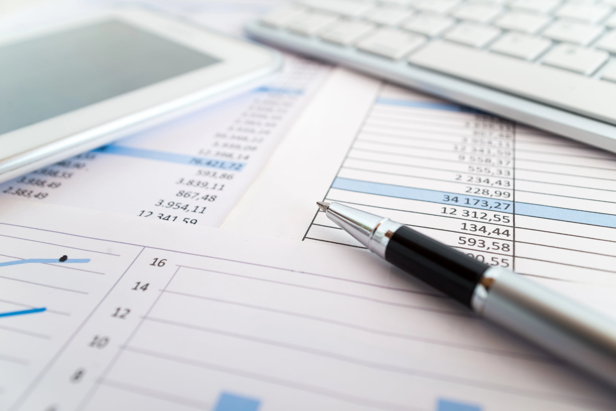 Close-up of a pen on a printed financial spreadsheet with a calculator and keyboard blurred in the background.